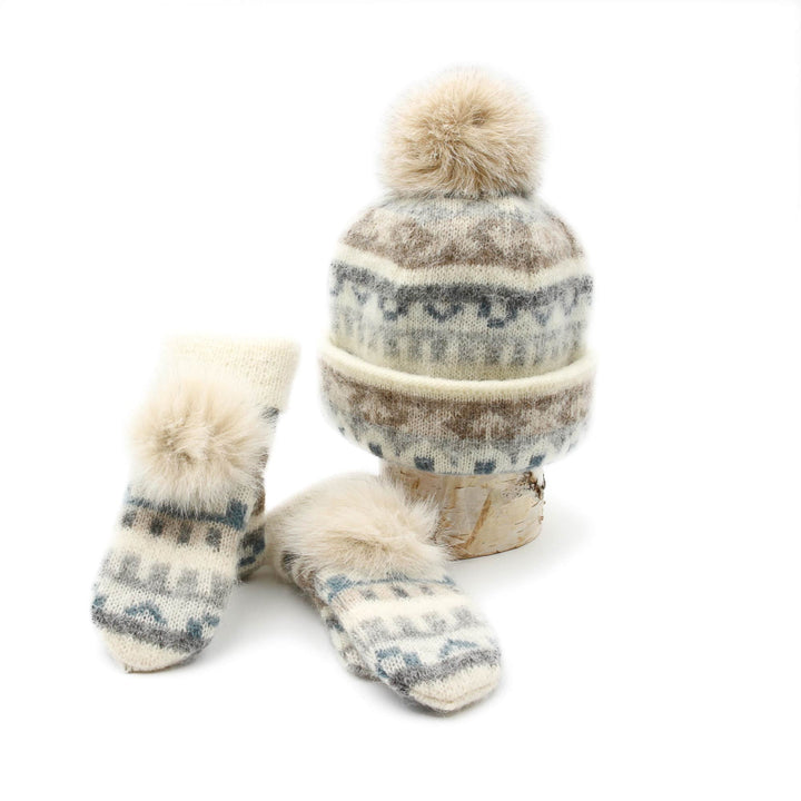 Mitaines et tuque en laine islandaises crèmes, grises et bleues avec pompom crème sur le dessus déposées contre une buche