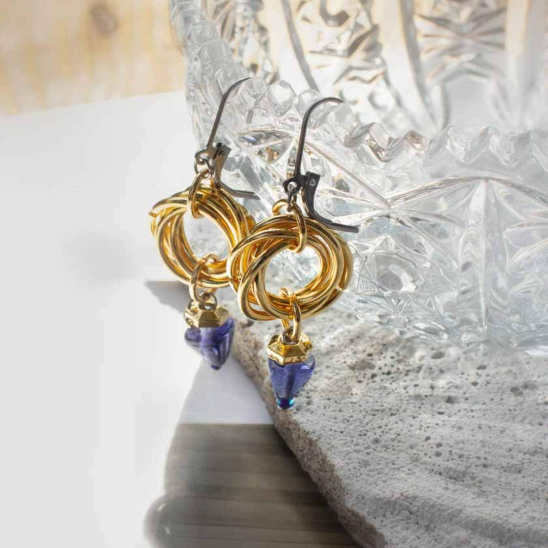 Boucles d'oreilles Anne-Marie Chagnon Jilka avec crochets argentés, anneaux dorés et pierre triangulaire mauve suspendue déposées contre un bol