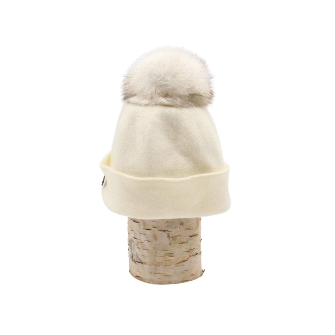Tuque Odetta blanche par Canadian hat avec un pompom blanc sur le dessus déposée sur une bûche