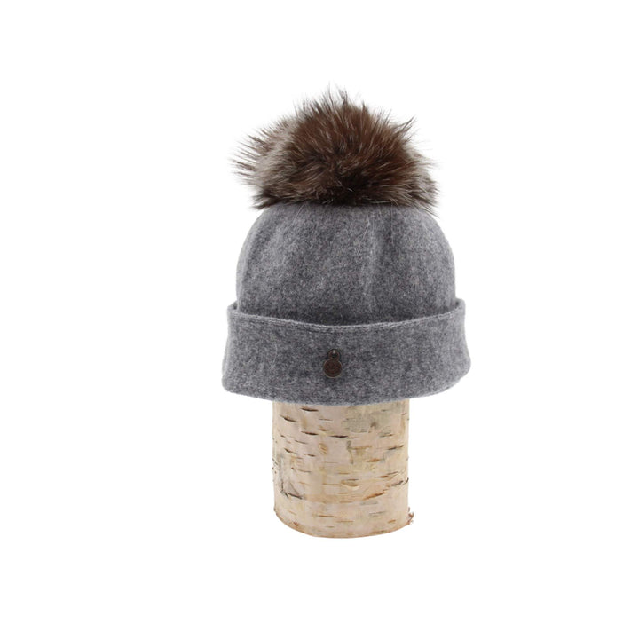 Tuque Odetta grise par Canadian hat avec un pompom brun sur le dessus déposée sur une bûche vue de côté