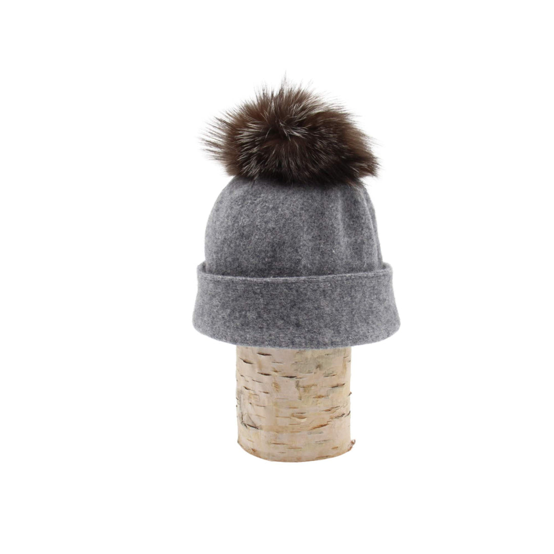 Tuque Odetta grise par Canadian hat avec un pompom brun sur le dessus déposée sur une bûche