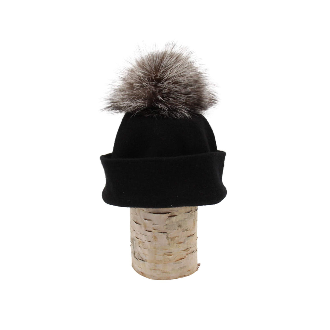 Tuque Odetta noire par Canadian hat avec un pompom gris sur le dessus déposée sur une bûche