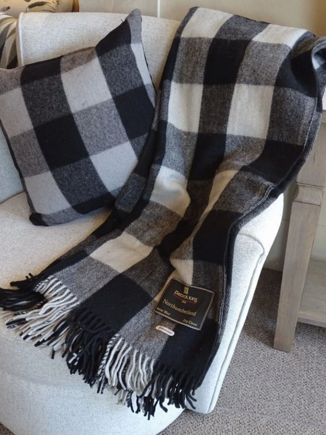 Lourde couverture tartan Northumberland noire et grise déposée à droite d'un coussin tartan assorti sur un fauteuil blanc