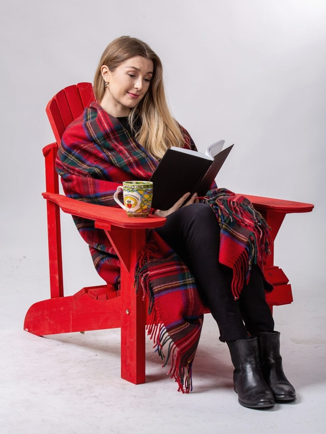 Lourde couverture tartan Highland Collection rouge avec lignes vertes,blanches, noires et bleues sur une femme lisant un livre sur une chaise rouge avec une tasse jaune