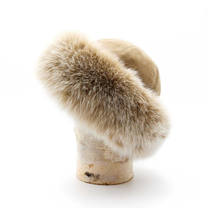chapeau de fourrure de renard style cloche posé sur une buche de bouleau. Le cuir suède est de couleur beige et la fourrure de renard est beige pâle.