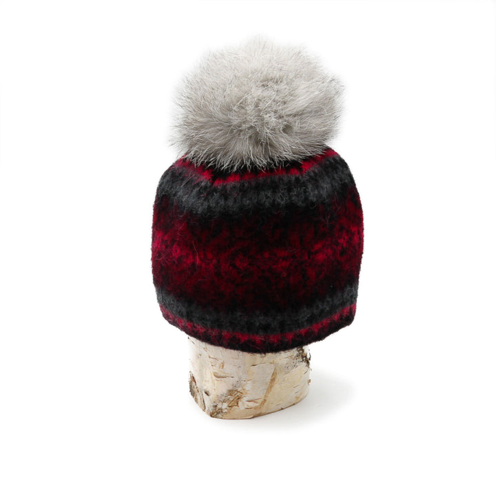 Tuque en laine islandaise noire,rouge et grise avec un pompom gris déposée sur une buche