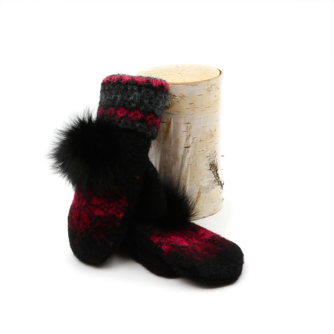 Mitaines en laine islandaise noires, rouges et grises avec pompom noir sur le dessus déposées contre une buche